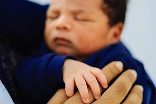 Photo of a young infant sleeping in an adult’s arms, their tiny hand holding the adult’s finger.