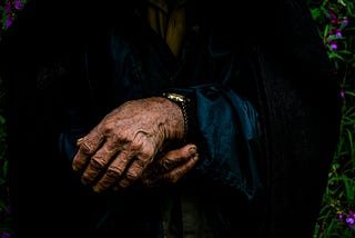 Close-up of an elderly person’s folded hands, worn with age, resting gently against a dark coat — evoking memory, time, and loss.