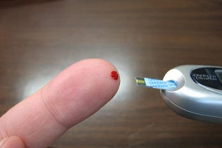 A finger with a drop of blood on the tip, adjacent to a blood sugar test strip.