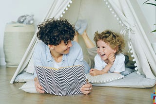 An adult reading and laughing with a small child in an indoor tent