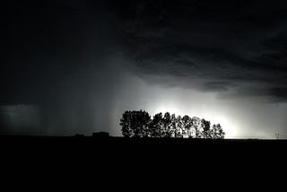 rain clouds hover over trees at night- you can see some light from behind the trees