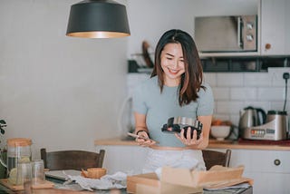 A young Asian woman unboxing headphones as she holds her cell phone.