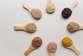 Eight small wooden spoons, each filled with various colors of protein powder, face with their heads inward to form a circle.