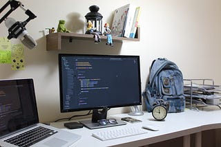 Computer desk featuring a computer monitor, backpack, alarm clock, and laptop.