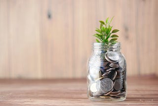 A small green plant growing out of a mason jar will with coins