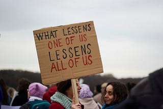 Woman holding a sign that says “What Lessens One of Us Lessens All of Us”