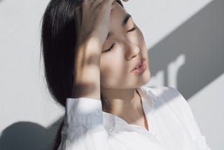 Fair-skinned young woman with neck-length straight, black hair and wearing long-sleeved white blouse. Her partially shadowed face, with eyes closed and lips partly opened, is upturned slightly sideways to the right, her right arm resting on a counter top and right hand on her forehead, as if massaging it.