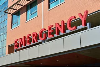 This is a stock photo that displays the front of a brick hospital building with the word “Emergency” in red capital letters above the entrance.