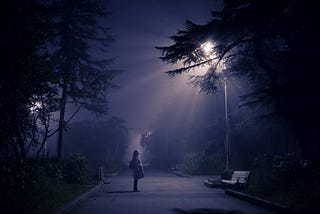 Photo of a woman standing in the middle of a road staring up at a streetlamp
