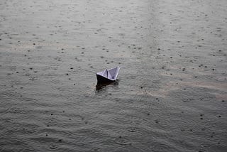 Photo of raindrops falling on water while a small paper boat bobs up and down.