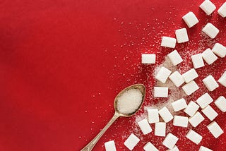 Sugar cubes scattered on a red background alongside a teaspoon of sugar.