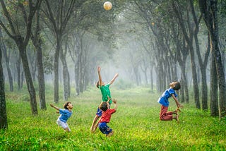 Four boys in colorful red, green, dark blue and pale blue T-shirts and shorts trying to catch a white ball high over their heads , in a grassy field with a line of tall green trees on both sides of the field that fades into a foggy background.