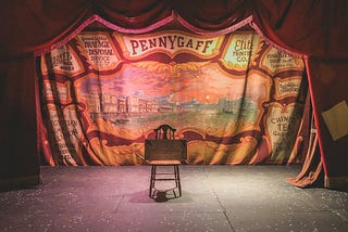 A stage, surrounded by a heavy, old-fashioned, red and gold entrance, is lit up to show a chair, used as an easel, holding a blank wooden board.