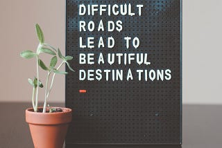 Little green plant next to black and white sign which reads “Difficult roads lead to beautiful destinations”