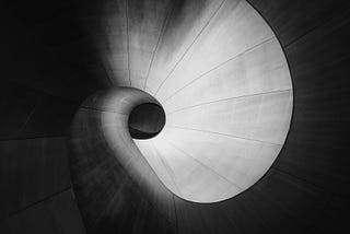 A high-contrast, black-and-white architectural photograph looking down into a smooth, sweeping spiral vortex, representing the complex and deep neural pathways of human memory.