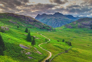 A path winds through a green, verdant valley, pointing to mountains in the distance