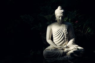 At night, a statue of what is probably understood to be the Buddha, tucked away in a garden, illuminated by light from a source which might be the moon.