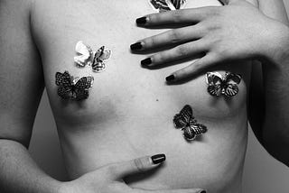 A naked chest of a person with butterfly pasties, and their hands with painted nails touching their body.