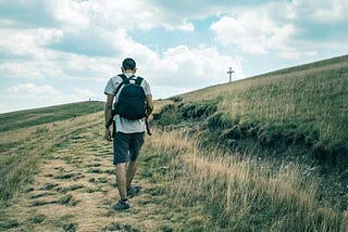 Man in white tee shirt and black shorts carrying a black back pack walking up a dry, barren hill toward a cross on a hill. The author is using the picture to show that having faith in the way to get somewhere is not always clear, but to follow God’s guidance and you will arrive.