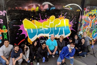 A group in front of a multi-colored mural with the words “Design” painted on a wall.