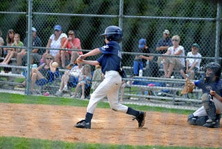 Young baseball player up to bat mid swing with a catcher behind him. A crowd watches him from the bleachers.