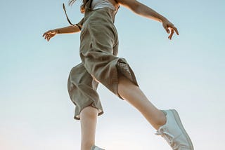 This image captures a woman in mid-air as she leaps or jumps, giving the appearance of floating against a clear sky. Her movement is dynamic, with her hair flowing wildly and her limbs gracefully extended. She wears a light, sleeveless jumpsuit and white sneakers, which contrast with the soft blue of the sky in the background. The image evokes a sense of freedom and weightlessness.
