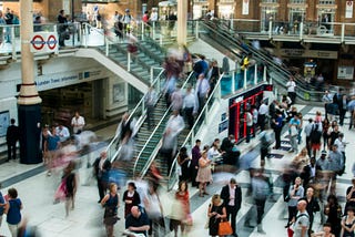 Wide angle view of a train station with multiple escalators and sets of stairs and many people. There are some shop fronts visible in the picture. Some of the people are blurry with movement.
