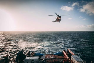 A helicopter flys above the ocean following behind the stern of a vessel