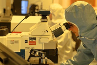 Lab technician, fully protected from the head down, working on a microscope in a big lab, other technicians behind