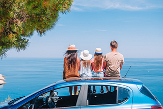 Woman, man, and two young girls sitting on top of a small car, looking out over the ocean.