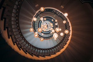 Looking up at a spiral staircase with many light bulbs hanging in the middle.