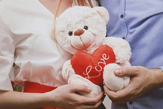 A couple standing close together, holding a white teddy bear with a red heart that reads “love,” their hands gently touching around the plush toy.