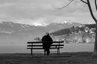 Old man alone on a park bench beside a lake with mountains in the background