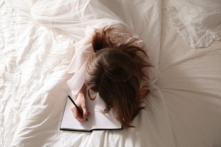 A woman lays in white sheets, writing a letter.