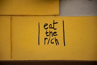 “Eat the rich” written in black marker on a yellow wall.