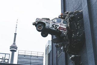 A picture of a truck that has smashed into the second story of a building and is suspended in the air with its front end breaking through the brick of the structure.