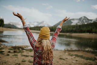 A woman in flannel looks over a mountain landscape with her arms in the air.