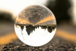 a glass ball outside on a road, looking through it’s illusion is an upside down view of some trees, the road, and yellow lines on it, etc.