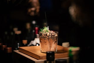 A mixed drink bubbling with a light green garnish in golden liquid and light — almost making it glow — sitting on a golden wooden board, elevated on a bartender’s counter but all else is dark around it except the silhouettes and shadows of liquor bottles.