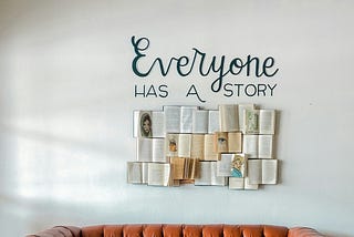 On a white wall above an orange sofa is a collage of books open with Everyone has a story written above it.