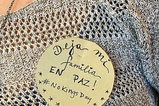 On a light yellow campaign-style button a barista write “Deja mi familia EN PAZ! #NoKingsDay”. Half of it is written in pretty cursive, the other half in all caps, followed by the hashtag for the October 18, 2025 march. She pinned it to her light gray crocheted sweater top.