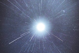 A bright, beautiful, burning, blue and white light shines in the night sky. There appears to be white, thin, beams of light shooting by around it.