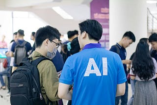 Two men with their backs to us. One is wearing a rucksack and a Covid-style mask. He is listening to the other, who is wearing a T-shirt with ‘AI’ printed on the back.