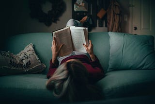 A woman lays on a blue-green sofa reading a book, so that we can see the pages.