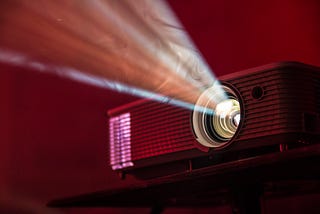 An LED projector sitting on a table. The projector is on and emitting light, which shines through dust.
