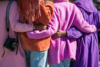 The backs of four Black women in brightly colored coats. They each have their arms around one another.