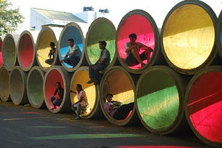 6 people casually sitting inside large cement pipes stacked in two rows, and covered on one opening side with blue, green, red, yellow film — to let filtered coloured light pass