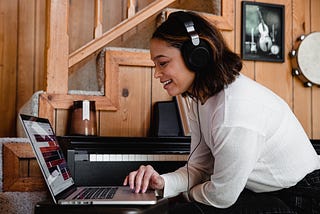 A woman with her headphones on, smiling and looking at the screen of the laptop.