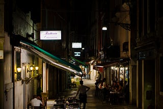 A nighttime alley lined with awnings of stores and cafes. Down the middle of the alley, one waitress with hand on hip.