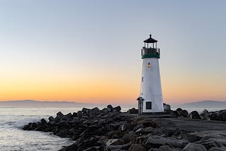 Photo of a lighthouse on a base of rocks, with a misty background and horizon line that suggests it is sunset